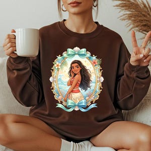 May include: A brown sweatshirt featuring a colorful oval graphic of a cartoon woman with long dark hair, wearing a red and white top and a white skirt. The graphic is framed by a blue ribbon, flowers, and seashells. The woman is holding a white mug.
