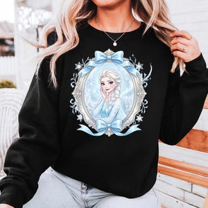 K&ouml;nnte beinhalten: Schwarzes Sweatshirt mit einem gerahmten Bild einer Prinzessin in einem blauen Kleid und blondem Zopf. Der Rahmen ist mit Schneeflocken und blauen Schleifen verziert. Die Prinzessin l&auml;chelt.