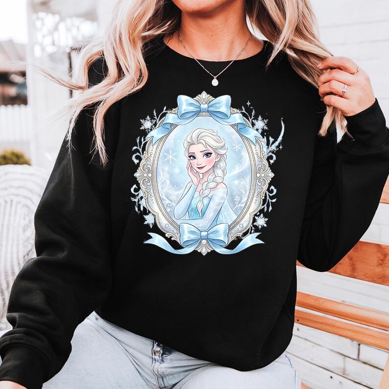 K&ouml;nnte beinhalten: Schwarzes Sweatshirt mit einem gerahmten Bild einer Prinzessin in einem blauen Kleid und blondem Zopf. Der Rahmen ist mit Schneeflocken und blauen Schleifen verziert. Die Prinzessin l&auml;chelt.