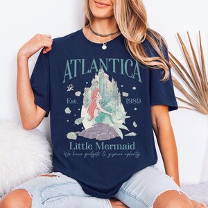 May include: Navy blue t-shirt featuring a graphic of a mermaid with the text "Atlantica" and "Little Mermaid". The design includes a castle, seashells, and the year "1989". The shirt has short sleeves and a crew neck.