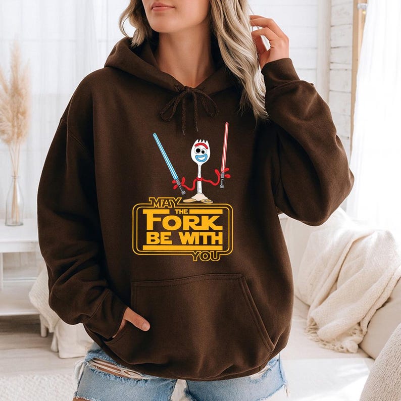 May include: A dark brown hooded sweatshirt featuring a graphic of a white plastic spork character with red pipe cleaner arms holding a blue and a red lightsaber. Below the character, yellow text reads: MAY THE FORK BE WITH YOU.