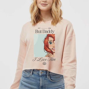 May include: A light pink cropped sweatshirt with the text "But Daddy" above an illustration of a cartoon character with red hair. The text "I Love Him" is below the illustration.