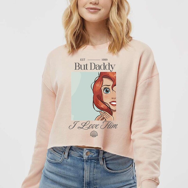 May include: A light pink cropped sweatshirt with the text "But Daddy" above an illustration of a cartoon character with red hair. The text "I Love Him" is below the illustration.
