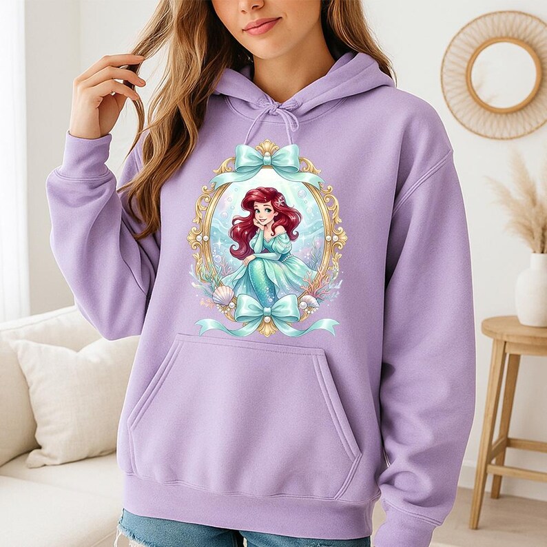 Puede incluir: Una sudadera con capucha lila con un bolsillo delantero y una capucha con cord&oacute;n. El dise&ntilde;o incluye una ilustraci&oacute;n enmarcada de una sirena con cabello rojo y un vestido turquesa, rodeada de conchas marinas y plantas acu&aacute;ticas. Lazos turquesas adornan el marco.