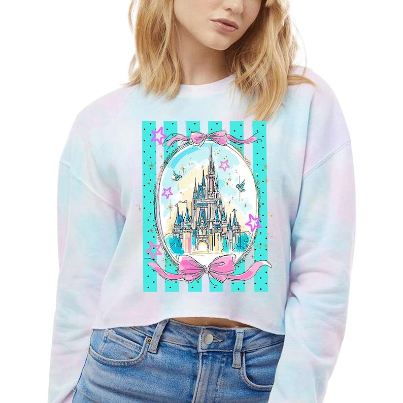 May include: White cropped sweatshirt with a tie-dye effect. The front features a watercolor illustration of a castle within an oval frame, accented with pink bows, stars, and a teal striped background.