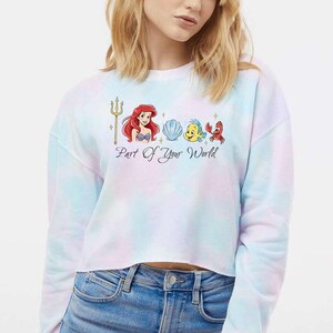 May include: A cropped tie-dye sweatshirt with a graphic of Ariel, Flounder, Sebastian, and a seashell. The text "Part Of Your World" is printed below the characters. The sweatshirt has a pastel colour scheme of blue, pink, and white.