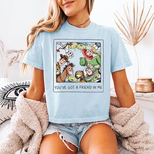 May include: Pale blue t-shirt with a graphic of characters from the animated film "Toy Story" within a square. The text "YOU'VE GOT A FRIEND IN ME" is printed below. The shirt is short-sleeved with a crew neck.