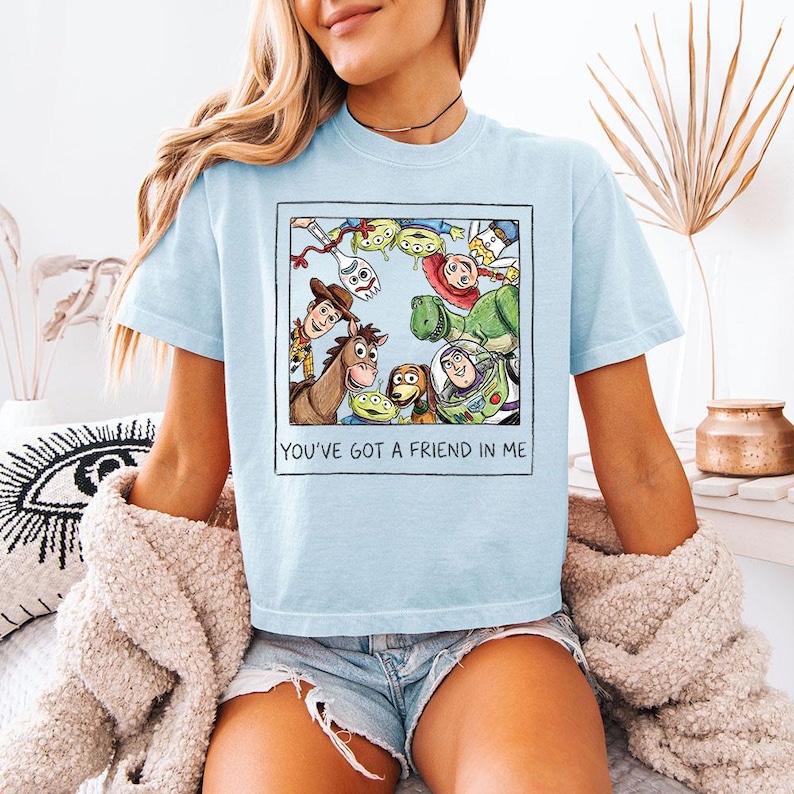 May include: Pale blue t-shirt with a graphic of characters from the animated film "Toy Story" within a square. The text "YOU'VE GOT A FRIEND IN ME" is printed below. The shirt is short-sleeved with a crew neck.