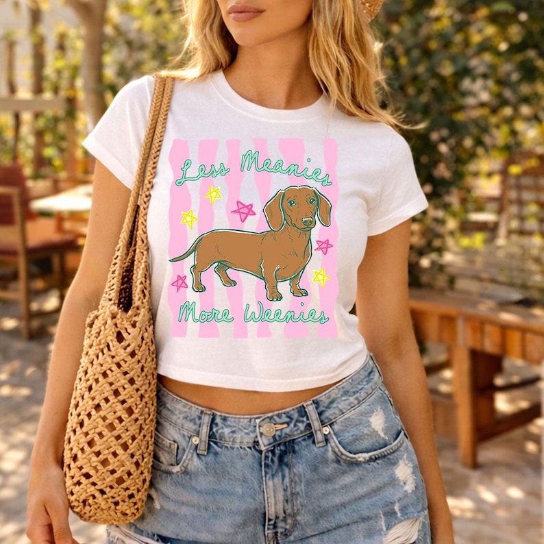 May include: A white short-sleeved t-shirt featuring a brown dachshund illustration, pink vertical wavy stripes, and colourful star accents. The text reads Less Meanies More Weenies in a green script font. The shirt is paired with a woven tan tote bag.