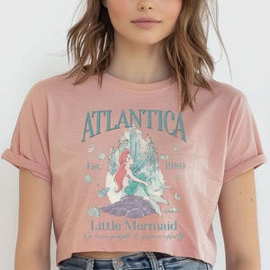 May include: A dusty rose crop top featuring a graphic of a mermaid and the text "Atlantica" and "Little Mermaid". The design includes a castle, seashells, and the year "1989". The shirt has rolled-up sleeves.