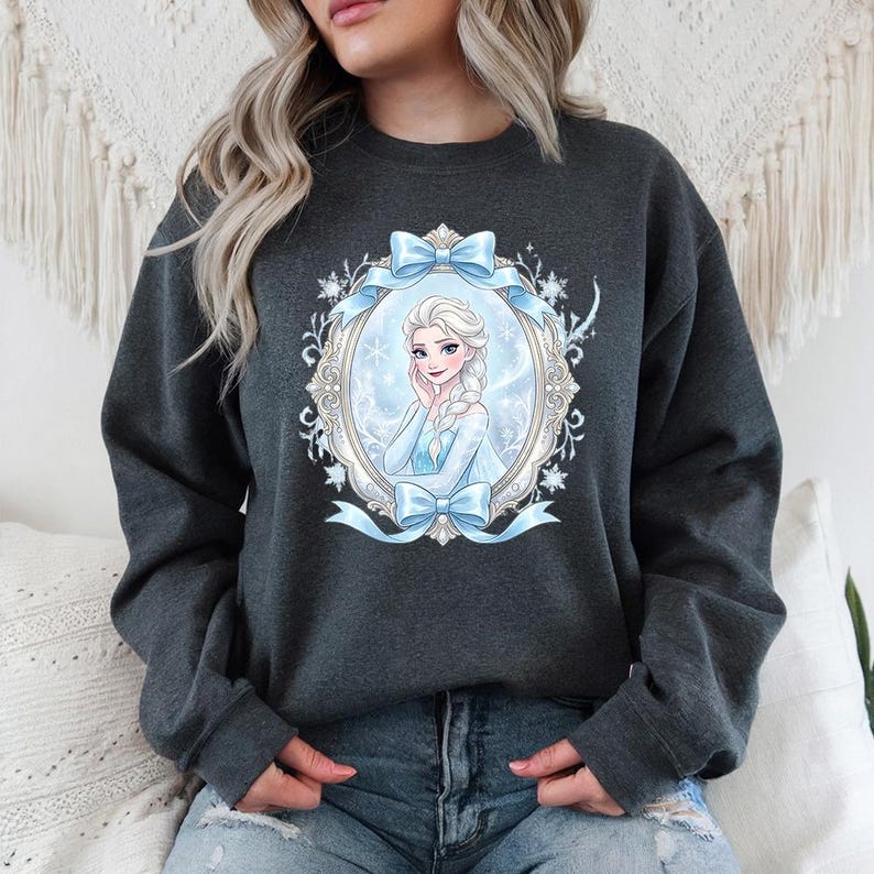 K&ouml;nnte beinhalten: Dunkelgraues Sweatshirt mit einem Cartoon-Portr&auml;t in einem ovalen Rahmen, verziert mit hellblauen Schleifen. Die Figur hat blondes Haar und tr&auml;gt ein hellblaues Kleid. Der Rahmen hat einen wei&szlig;en und goldenen Rand.