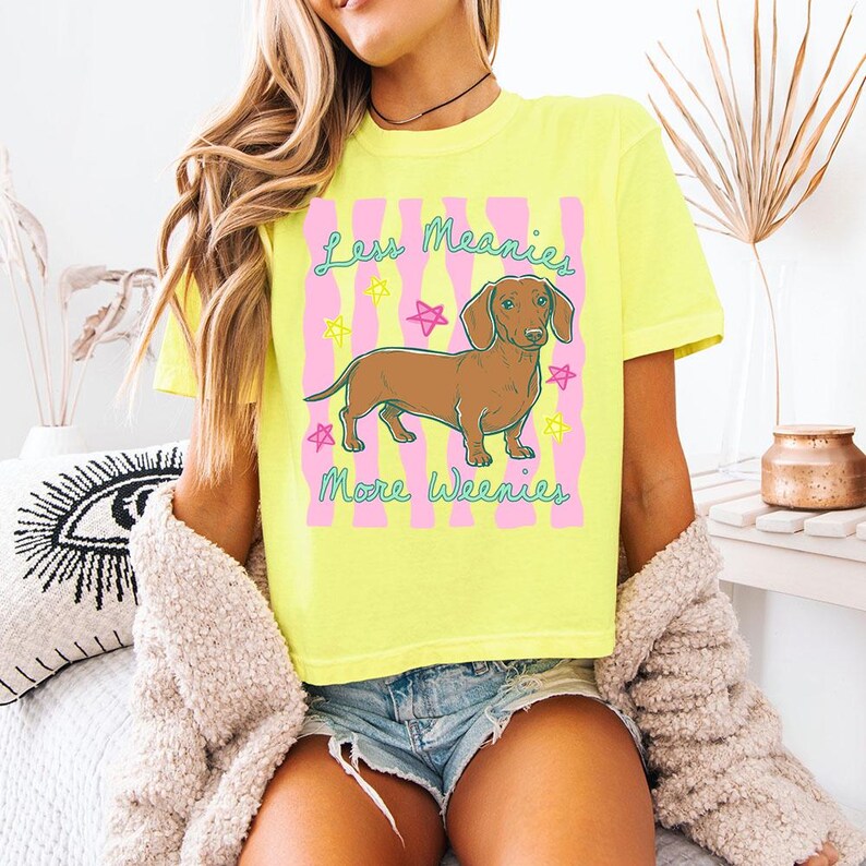 May include: A neon yellow short-sleeve t-shirt featuring a graphic of a brown dachshund dog. The design includes pink vertical wavy stripes, colourful stars, and the text "Less Meanies More Weenies" in a teal cursive font across the front.