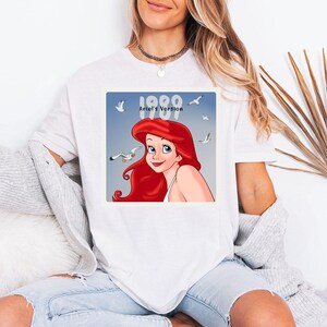 May include: White t-shirt featuring a graphic of Ariel from The Little Mermaid. The graphic includes the text "1989 Ariel's Version" with seagulls in the background. The cartoon Ariel has red hair and a smiling expression.