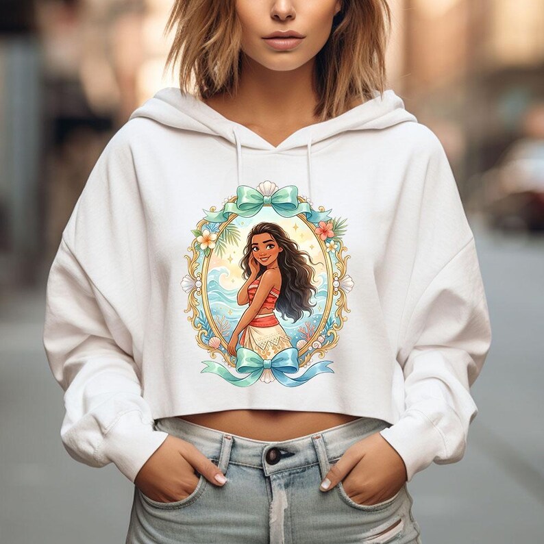 K&ouml;nnte beinhalten: Wei&szlig;er, gecroppter Kapuzenpullover mit einer farbenfrohen Illustration einer Frau in tropischer Umgebung. Das Kunstwerk ist von einem ovalen Rahmen mit floralen und Schleifenakzenten umgeben. Der Hoodie hat lange &Auml;rmel und eine Kapuze mit Kordelzug.