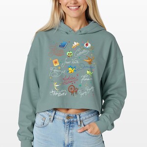 May include: A sage green cropped hoodie featuring a graphic print of various animated character signatures, including Ariel, Cinderella, and Jasmine, alongside small colorful icons like a golden lamp, a spinning wheel, and a blue book.