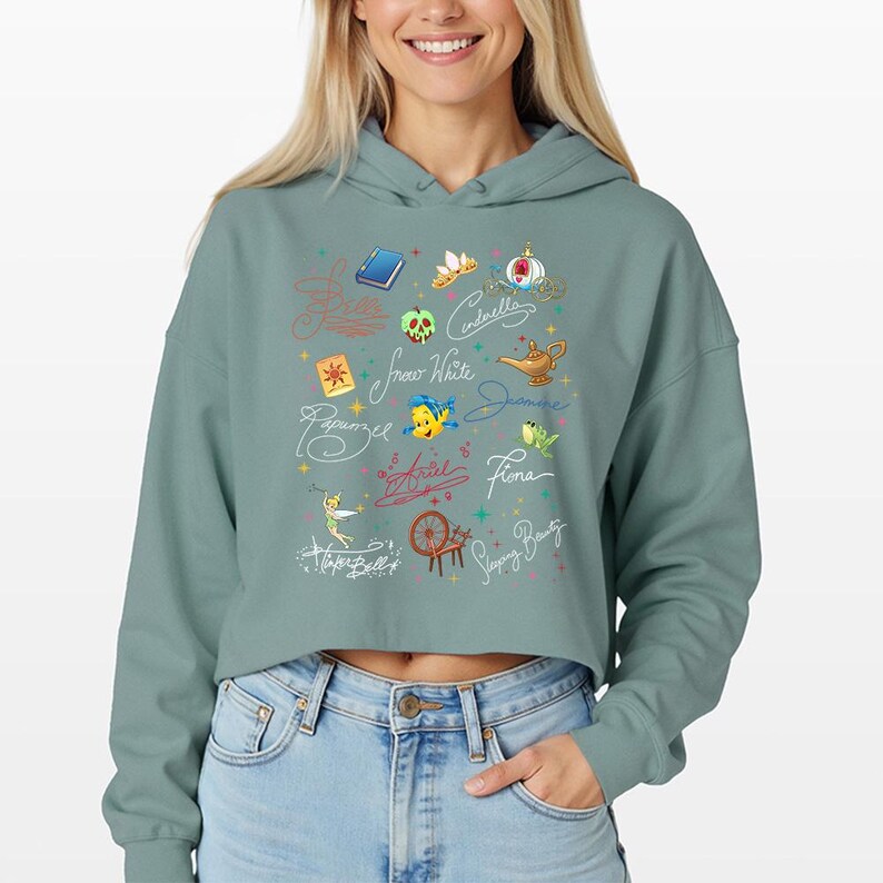 May include: A sage green cropped hoodie featuring a graphic print of various animated character signatures, including Ariel, Cinderella, and Jasmine, alongside small colorful icons like a golden lamp, a spinning wheel, and a blue book.