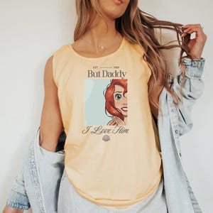 May include: A light yellow tank top with the text "But Daddy I Love Him" and an illustration of a red-haired character. The text "EST. 1989" is above the text. A seashell graphic is below the text.