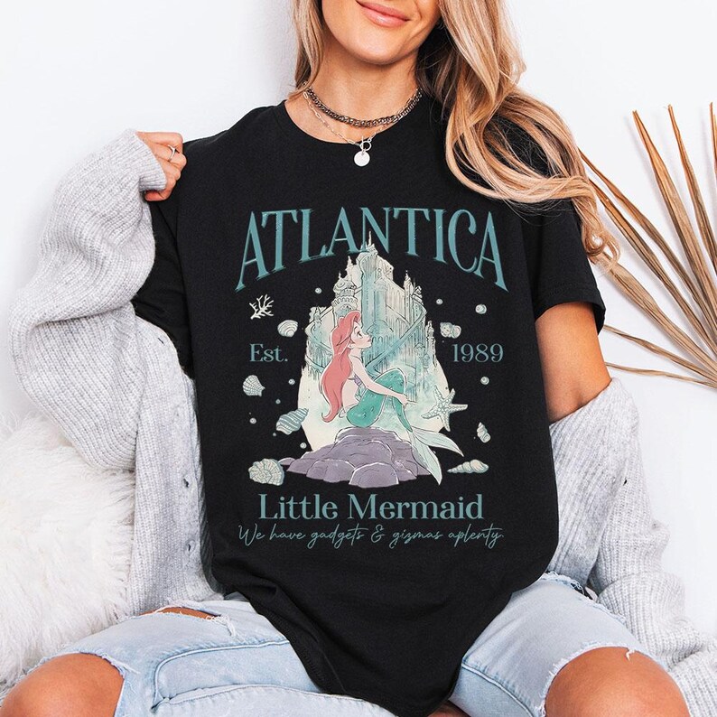 May include: Black t-shirt featuring a teal graphic with the word "Atlantica" above an illustration of a mermaid and castle. The text "Est. 1989" and "Little Mermaid" are also included. The phrase "We have gadgets & gizmos aplenty" is printed below.