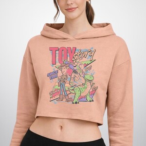 May include: A peach-coloured cropped hoodie featuring a graphic print of Toy Story characters Woody, Buzz Lightyear, and Rex. The design includes the text "TOY story", "SINCE 1995", "Woody", "Buzz", and "Rex" in a colourful, retro-inspired artistic style.