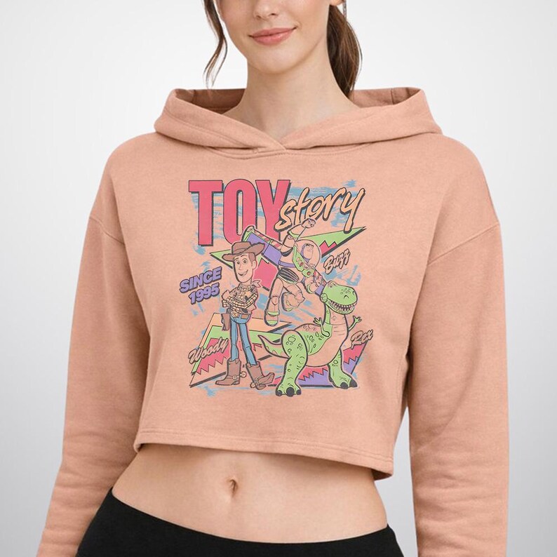 May include: A peach-coloured cropped hoodie featuring a graphic print of Toy Story characters Woody, Buzz Lightyear, and Rex. The design includes the text "TOY story", "SINCE 1995", "Woody", "Buzz", and "Rex" in a colourful, retro-inspired artistic style.
