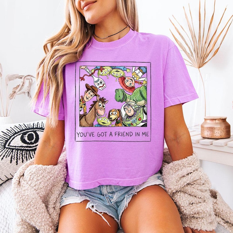 May include: A lilac t-shirt with a framed graphic of Toy Story characters, including Woody and Buzz Lightyear. The text "YOU'VE GOT A FRIEND IN ME" is printed below the graphic. The t-shirt is a short-sleeved, casual style.
