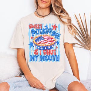 Op de afbeelding: Een beige t-shirt met korte mouwen en een afbeelding van een taart met een Amerikaanse vlag als korst. De tekst SWEET POTATO PIE & I SHUT MY MOUTH staat in blauwe en rode letters, omringd door blauwe verticale strepen en sterren.