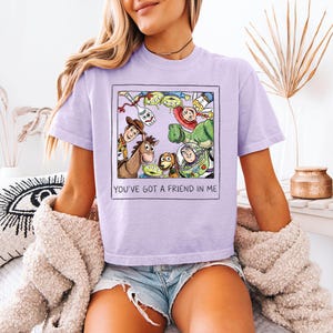 May include: A lilac t-shirt with a framed graphic of Toy Story characters, including Woody and Buzz Lightyear. The graphic is surrounded by a white border. The text "YOU'VE GOT A FRIEND IN ME" is printed below the graphic in black.