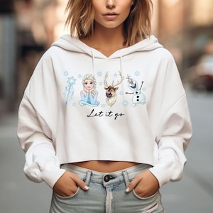 May include: White cropped hoodie with a graphic of Elsa, Sven, and Olaf from the film Frozen, snowflakes and the text "Let it go". The hoodie has a drawstring hood and long sleeves.