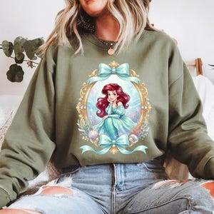 May include: An olive green sweatshirt featuring a framed illustration of a mermaid with red hair and a teal dress. The illustration is surrounded by teal bows and seashells. The sweatshirt is made of a soft material.