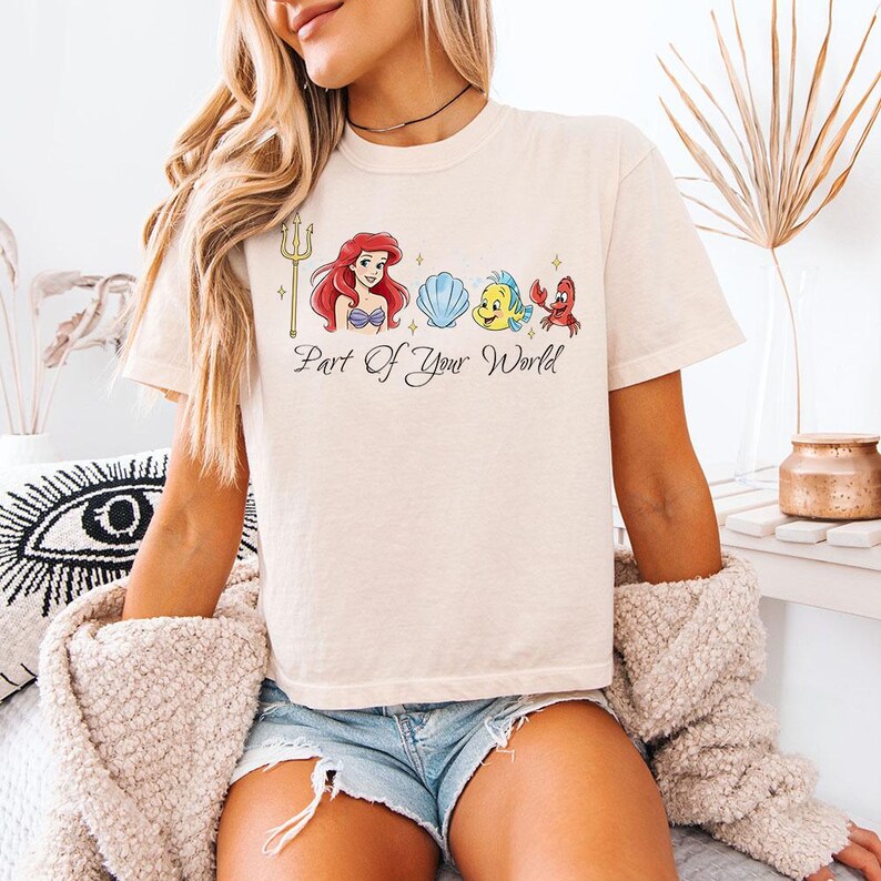 May include: Cream-colored t-shirt featuring a graphic of Ariel, Flounder, Sebastian, and a seashell. The phrase "Part Of Your World" is written below the characters. The shirt is a short-sleeved, casual style.