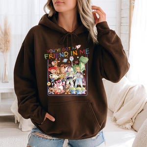 May include: A brown hooded sweatshirt featuring a colorful graphic of characters from the animated film "Toy Story." The graphic includes the text "YOU'VE GOT A FRIEND IN ME" in rainbow colors. The sweatshirt has a front pocket and a drawstring hood.