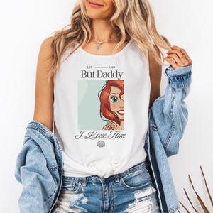 May include: White tank top featuring a graphic of a cartoon character with red hair and the text "But Daddy I Love Him." The design includes the year "1989" and a seashell graphic. The top is sleeveless.