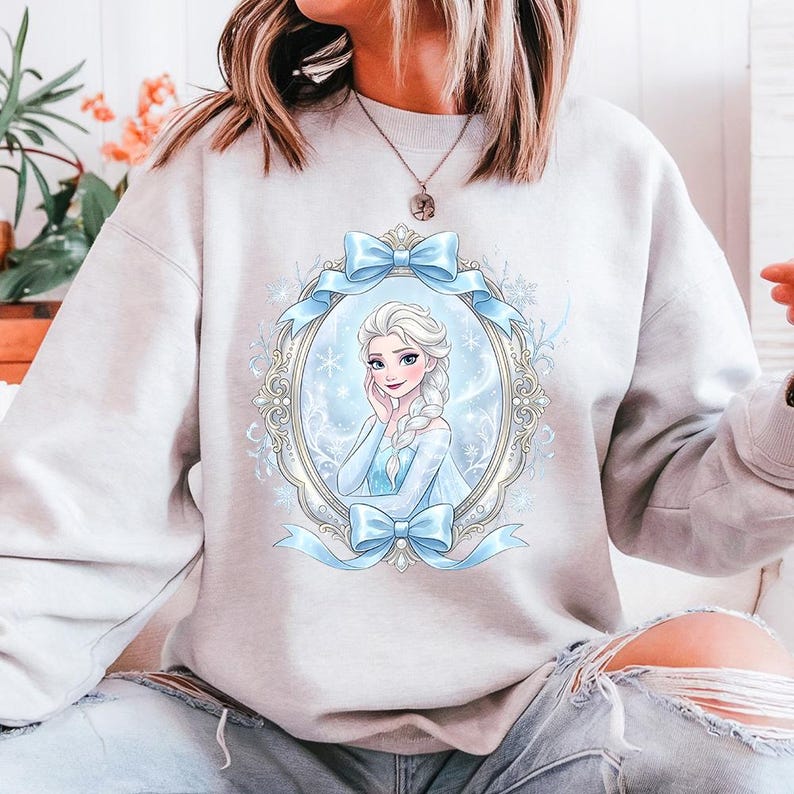 K&ouml;nnte beinhalten: Ein hellgraues Sweatshirt mit einer gerahmten Illustration einer Prinzessin in einem blauen Kleid und einem geflochtenen Haarschnitt. Der Rahmen ist mit Schneeflocken und blauen B&auml;ndern verziert. Das Sweatshirt ist ein l&auml;ssiger Stil.