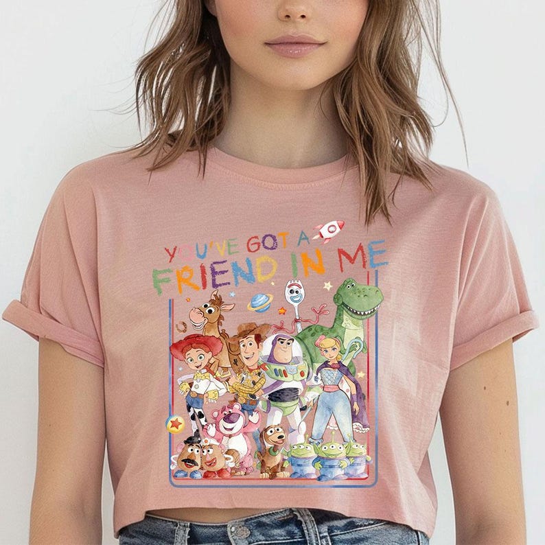 May include: A pale pink cropped t-shirt with a vibrant graphic of "Toy Story" characters. The design includes the phrase "YOU'VE GOT A FRIEND IN ME" in multiple colours. The top has short sleeves and a round neckline.