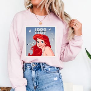 May include: Light pink sweatshirt featuring a graphic of Ariel from The Little Mermaid, with the text "1989 Ariel's Version." The illustration shows Ariel with red hair and blue eyes, surrounded by seagulls against a blue background.