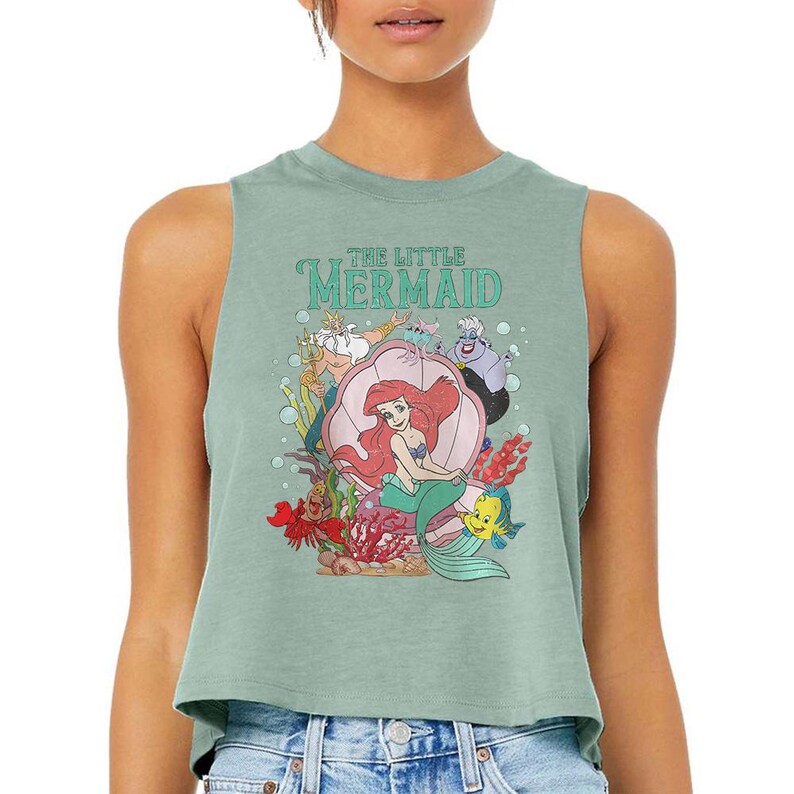 Puede incluir: Un top corto sin mangas de color verde salvia con un estampado gr&aacute;fico de los personajes de La Sirenita, incluyendo a Ariel, el Rey Trit&oacute;n, &Uacute;rsula, Sebasti&aacute;n y Flounder. El dise&ntilde;o incluye una gran concha rosa, plantas marinas y burbujas.