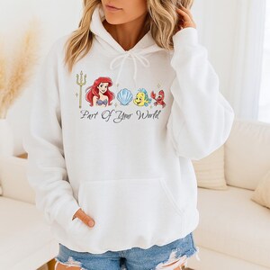 May include: White hooded sweatshirt featuring a colorful graphic of a trident, a mermaid, a seashell, a yellow fish, and a red crab. The text "Part of Your World" is below the graphic. The sweatshirt has a front pocket and a drawstring hood.