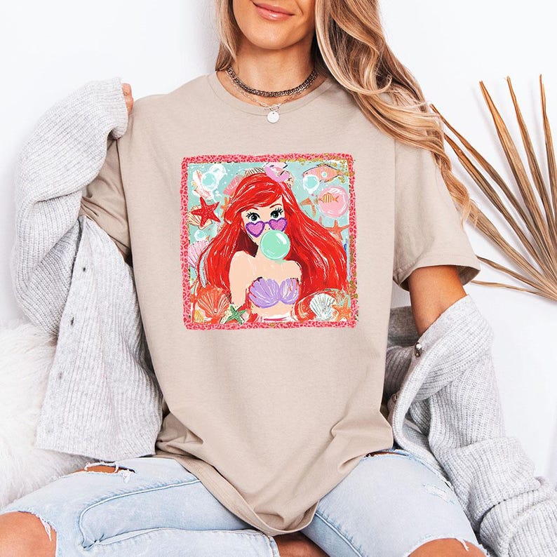 May include: Beige t-shirt featuring a colorful graphic of a mermaid with red hair, wearing heart-shaped sunglasses and blowing a bubble. The graphic includes seashells, starfish, and a pink border. The shirt is a casual, everyday wear item.