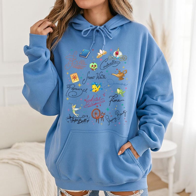 May include: A light blue hooded sweatshirt featuring a front graphic print of various animated character icons, including a magic lamp, a spinning wheel, a crown, and a poison apple, alongside handwritten names like Cinderella and Ariel.