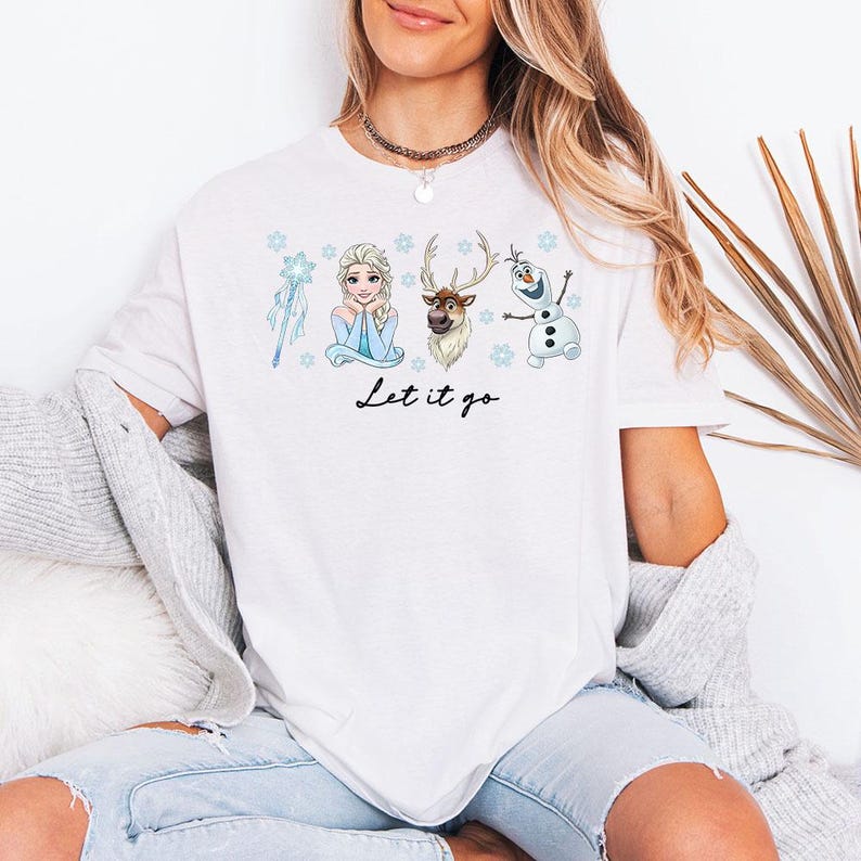 May include: White t-shirt with a colourful graphic of characters from the film Frozen, including Elsa, Olaf, and Sven, with the text "Let it go" in a script font. The design includes snowflakes and a magic wand.