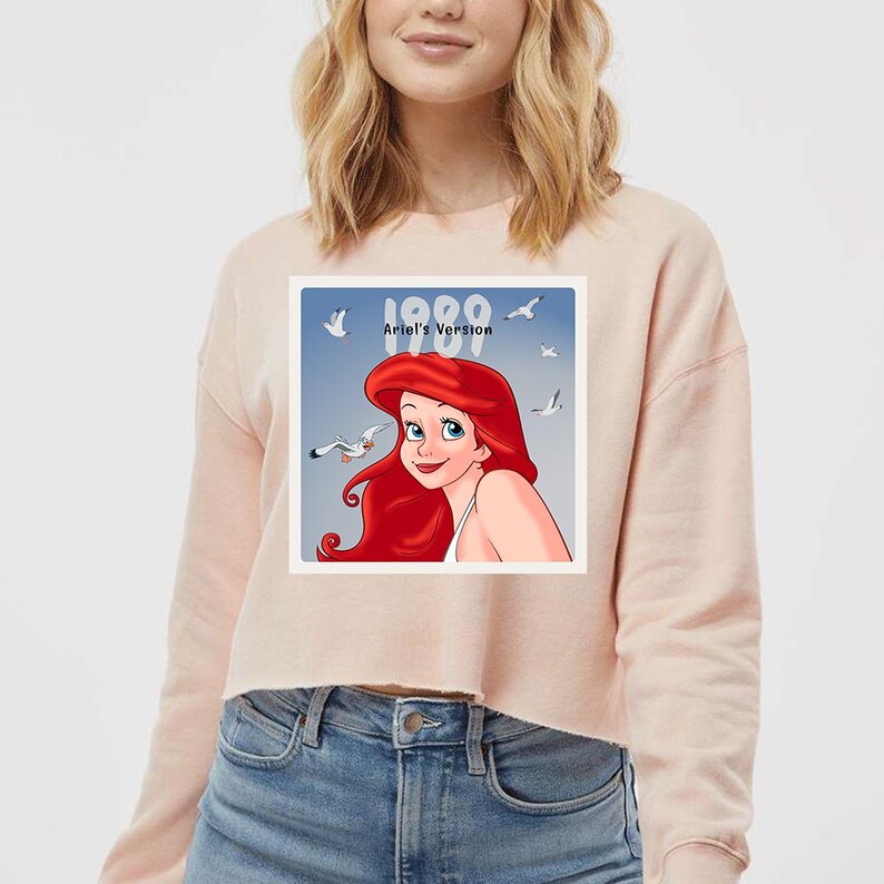Pode incluir: Um moletom cropped rosa claro com um gr&aacute;fico de Ariel de A Pequena Sereia. O gr&aacute;fico apresenta Ariel com cabelo ruivo e olhos azuis, com o texto "1989 Ariel's Version" em branco. O moletom &eacute; combinado com jeans azuis.