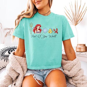 May include: A turquoise t-shirt featuring a graphic of Ariel, Flounder, Sebastian, and a seashell. The phrase "Part Of Your World" is printed below the characters. The shirt is a short-sleeved, crew neck style.