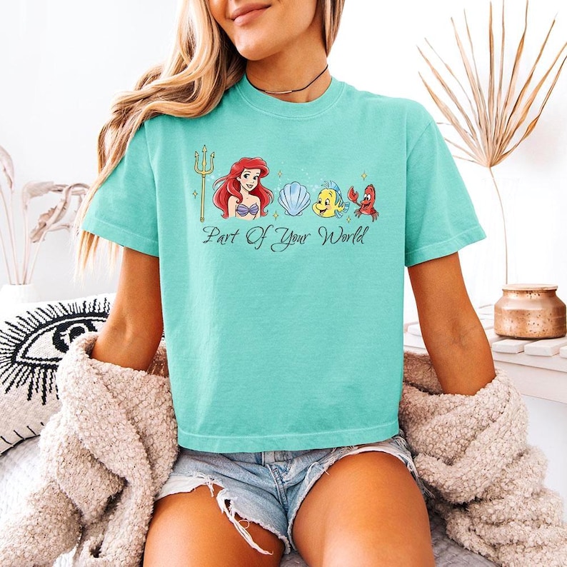 May include: A turquoise t-shirt featuring a graphic of Ariel, Flounder, Sebastian, and a seashell. The phrase "Part Of Your World" is printed below the characters. The shirt is a short-sleeved, crew neck style.