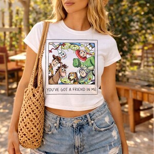 May include: White t-shirt with a colourful graphic of characters from the animated film "Toy Story." The design includes Woody, Buzz Lightyear, and other characters, with the text "YOU'VE GOT A FRIEND IN ME" below the image.