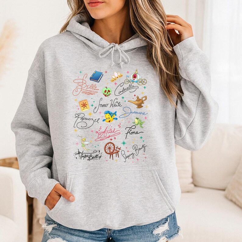 May include: A light gray hooded sweatshirt featuring a graphic print of various character names written in cursive, including Belle, Cinderella, Snow White, Jasmine, Rapunzel, Ariel, Fiona, Tinker Bell, and Sleeping Beauty, alongside small icons.