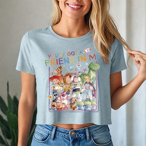 May include: Pale blue crop top with a vibrant graphic of Toy Story characters. The design includes the phrase "YOU'VE GOT A FRIEND IN ME" in rainbow lettering. The top is made from a soft fabric.