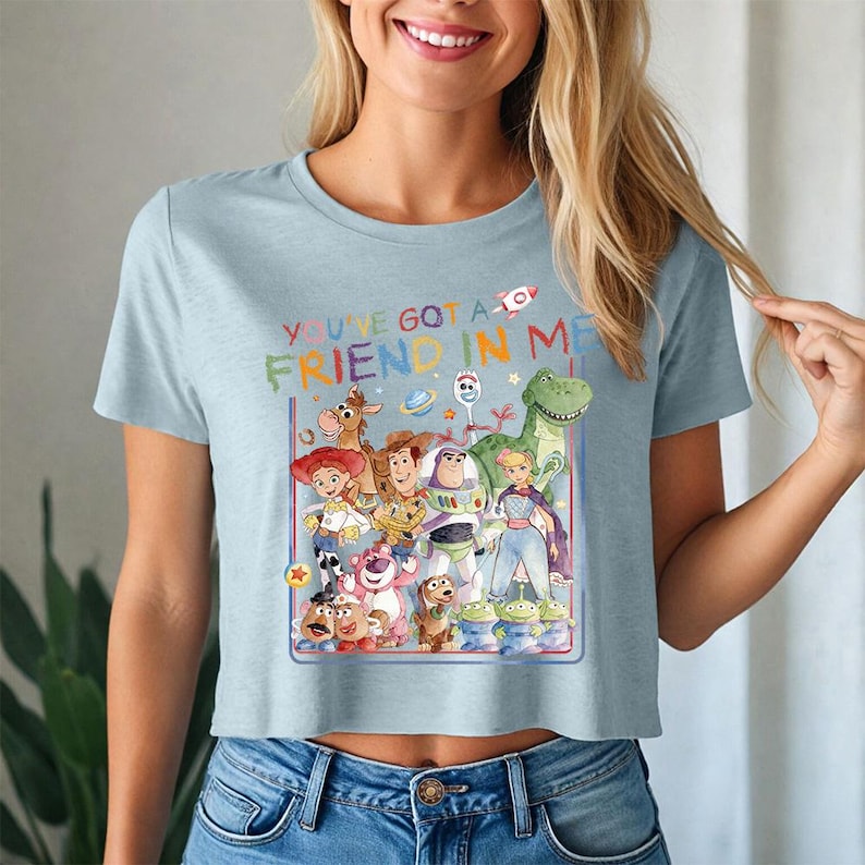 May include: Pale blue crop top with a vibrant graphic of Toy Story characters. The design includes the phrase "YOU'VE GOT A FRIEND IN ME" in rainbow lettering. The top is made from a soft fabric.