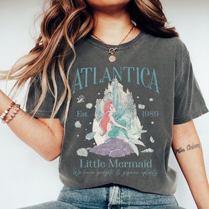 Op de afbeelding: Donkergrijs T-shirt met een afbeelding van een zeemeermin met rood haar en een turquoise staart. Het shirt heeft de tekst "Atlantica" boven de afbeelding en "Little Mermaid" eronder. Het shirt heeft ook de tekst "Est. 1989".