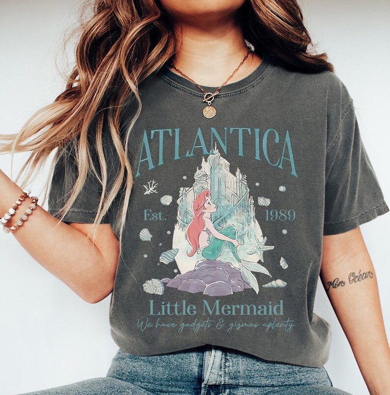 Op de afbeelding: Donkergrijs T-shirt met een afbeelding van een zeemeermin met rood haar en een turquoise staart. Het shirt heeft de tekst "Atlantica" boven de afbeelding en "Little Mermaid" eronder. Het shirt heeft ook de tekst "Est. 1989".