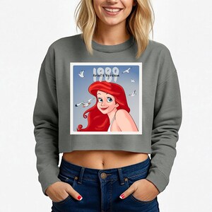 Pode incluir: Camisola cinzenta curta com um gr&aacute;fico de Ariel de A Pequena Sereia, com o texto "1989 Ariel's Version". O gr&aacute;fico mostra Ariel com cabelo ruivo e gaivotas contra um c&eacute;u azul. A camisola &eacute; de estilo casual.